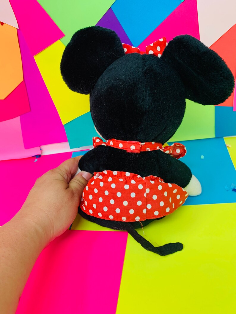 May include: A black and red plush toy mouse wearing a red polka dot dress and a red bow. The toy is sitting on a colorful background.