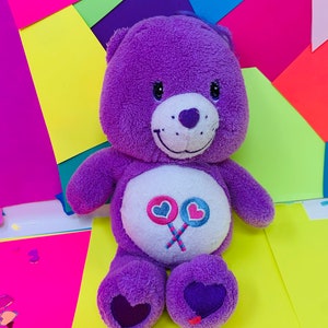 Pu&ograve; includere: Orso di peluche viola con pancia bianca e un design a lecca-lecca rosa e blu. L'orso ha un naso a forma di cuore ed &egrave; seduto su una superficie gialla.