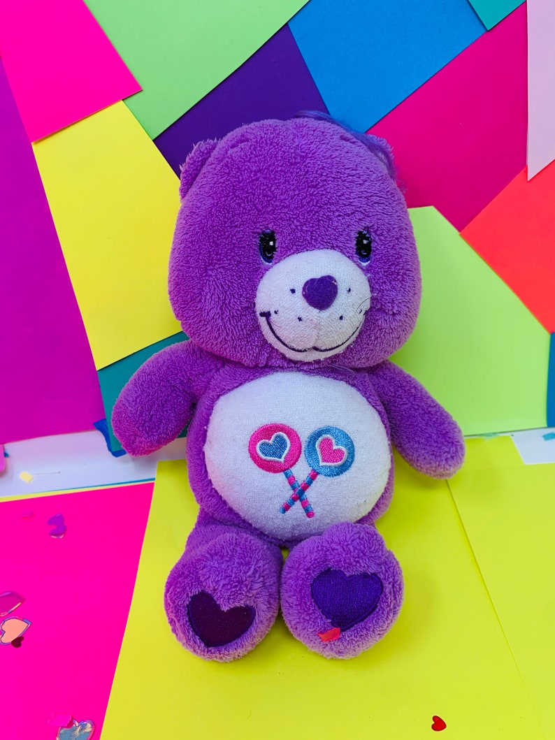Pu&ograve; includere: Orso di peluche viola con pancia bianca e un design a lecca-lecca rosa e blu. L'orso ha un naso a forma di cuore ed &egrave; seduto su una superficie gialla.