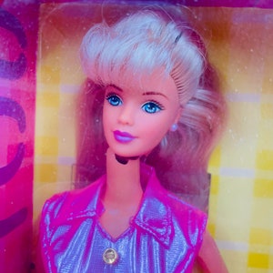 May include: A blonde Barbie doll wearing a pink and purple iridescent dress with a gold button. The doll has blue eyes and pink lips.