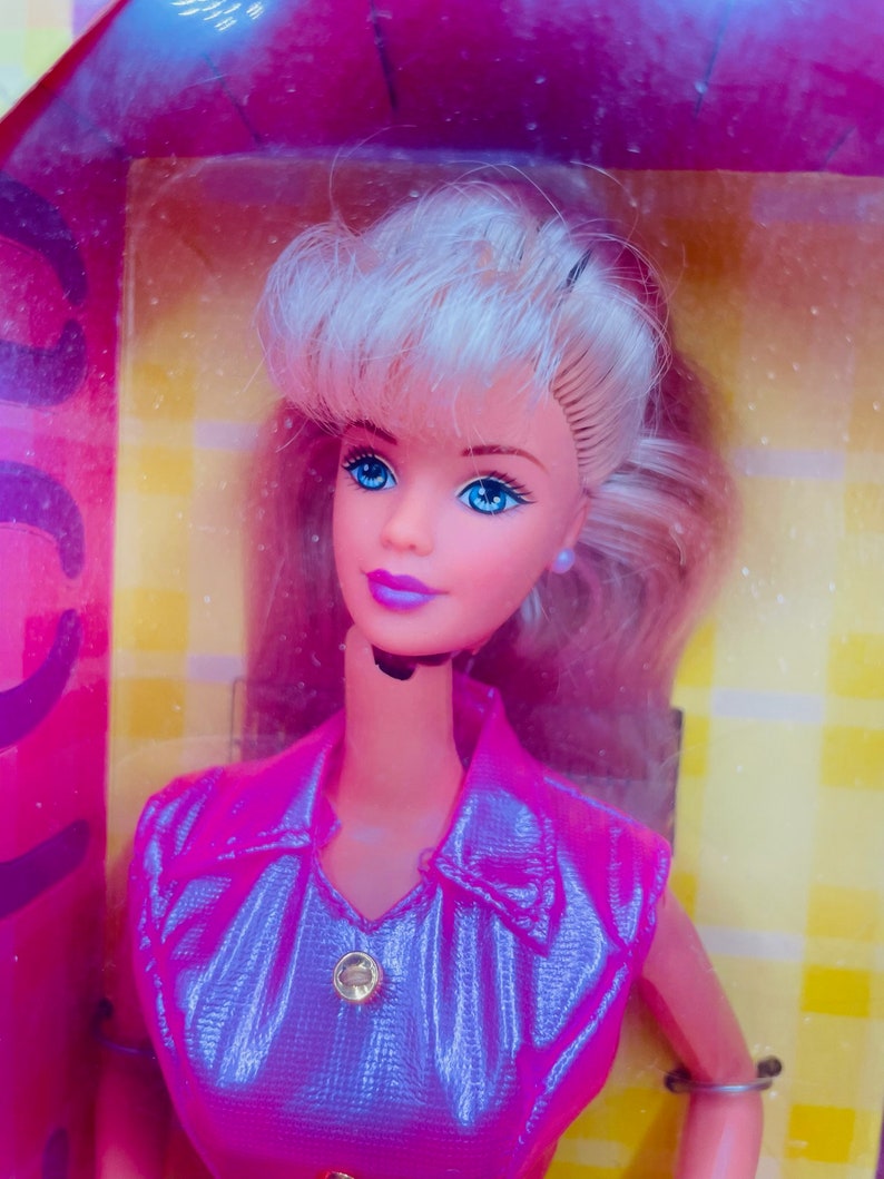 May include: A blonde Barbie doll wearing a pink and purple iridescent dress with a gold button. The doll has blue eyes and pink lips.