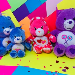 Pu&ograve; includere: Tre orsacchiotti Care Bears di peluche, uno blu, uno rosa e uno viola, sono seduti su uno sfondo colorato. Gli orsi blu hanno un cuore e un pianeta sul petto. L'orso rosa ha un cuore sul petto. L'orso viola ha un lecca-lecca con cuori.