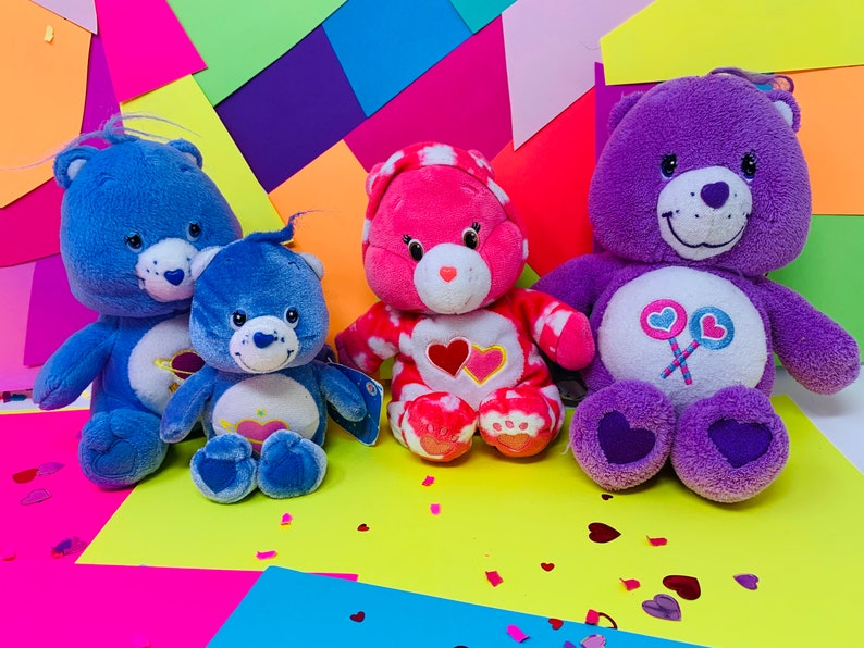 Pu&ograve; includere: Tre orsacchiotti Care Bears di peluche, uno blu, uno rosa e uno viola, sono seduti su uno sfondo colorato. Gli orsi blu hanno un cuore e un pianeta sul petto. L'orso rosa ha un cuore sul petto. L'orso viola ha un lecca-lecca con cuori.