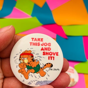 Könnte beinhalten: Ein weißer Knopf mit einer laufenden orangefarbenen Comic-Katze mit rotem Stirnband und grünem Badeanzug. Der Text auf dem Knopf lautet "Take this jog and shove it!" mit der Unterschrift "Jim Davis" unten.