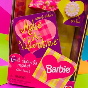 May include: A pink and yellow Barbie doll box with a heart-shaped design. The box says "Make a Valentine" and "Cool stencils inside!" with a Barbie logo. The box includes a pink and yellow plaid dress and a pink flower ring.