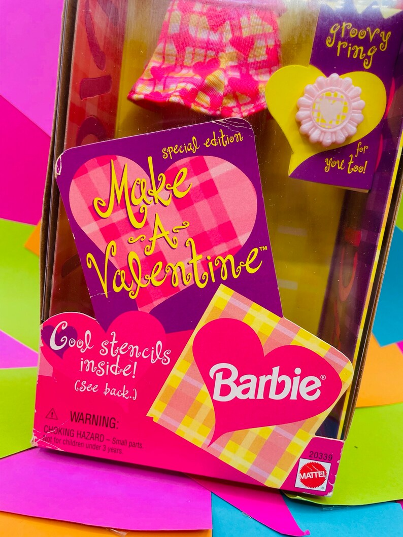 May include: A pink and yellow Barbie doll box with a heart-shaped design. The box says "Make a Valentine" and "Cool stencils inside!" with a Barbie logo. The box includes a pink and yellow plaid dress and a pink flower ring.