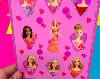 May include: A sheet of pink stickers featuring eight different images of Barbie dolls in various poses and outfits. Each sticker is surrounded by pink hearts and the text "Mattel" appears on each sticker.