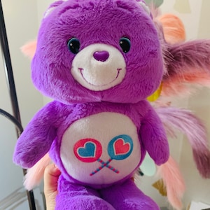 Pu&ograve; includere: Peluche di orso Care Bear viola con viso bianco e occhi blu. L'orso tiene due lecca-lecca rosa e blu con cuori. L'orso ha una coda rosa e bianca.