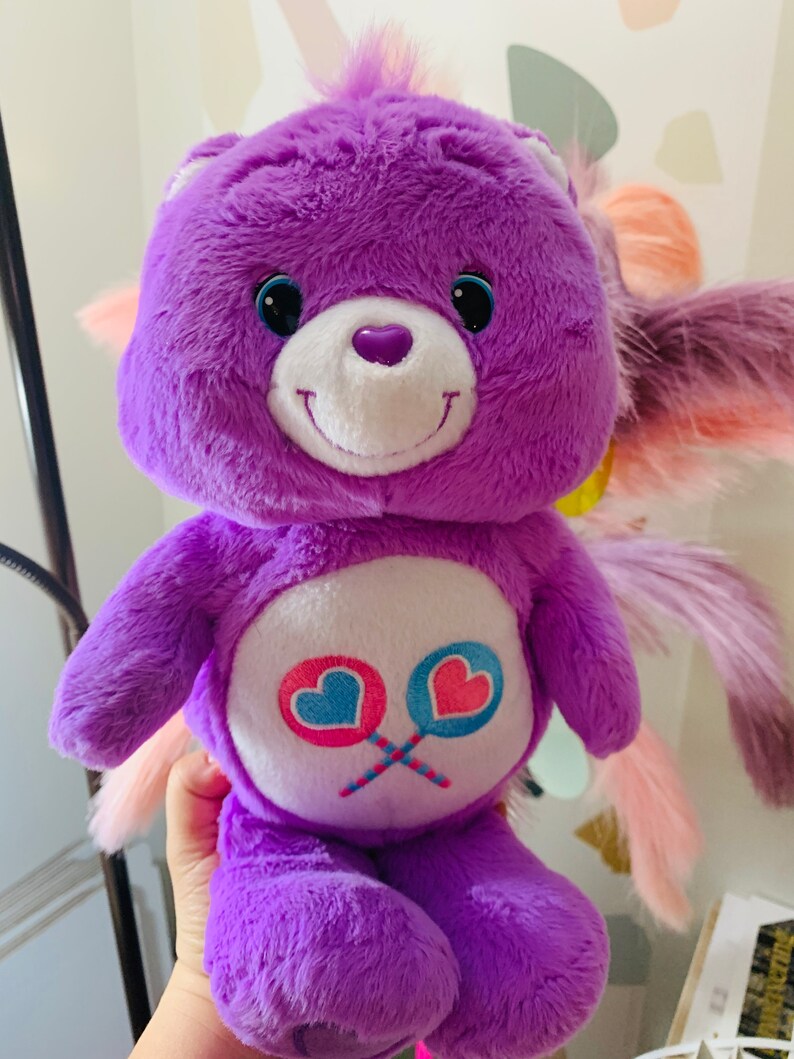 Pu&ograve; includere: Peluche di orso Care Bear viola con viso bianco e occhi blu. L'orso tiene due lecca-lecca rosa e blu con cuori. L'orso ha una coda rosa e bianca.