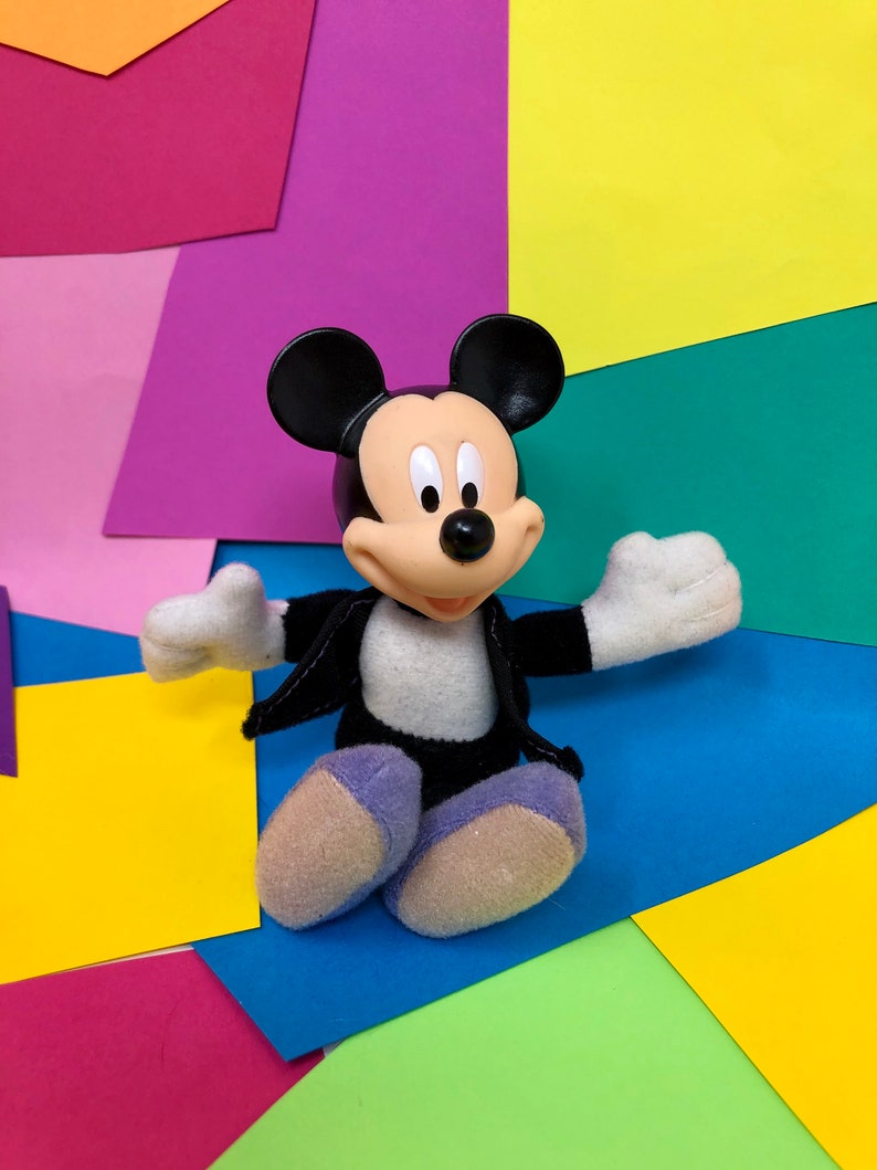 Op de afbeelding: Een zwart-wit Mickey Mouse knuffeldier met een zwart jack aan, zittend op een kleurrijke achtergrond van roze, paars, geel, groen en blauw papier.