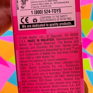 May include: Pink and white Barbie doll packaging with a pink background and a white border. The packaging features a photo of the Barbie doll and the text "YOU CAN TELL IT'S MATTEL!" and "MADE IN MALAYSIA".
