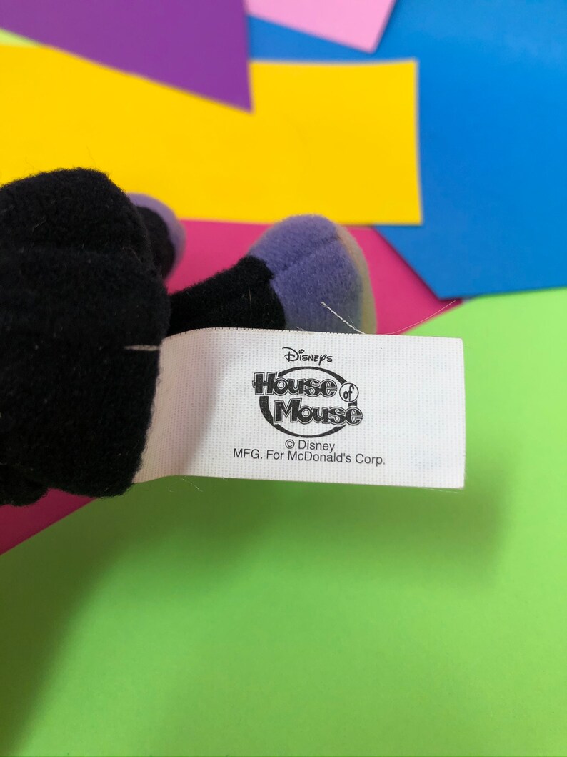 Op de afbeelding: Wit etiket met zwarte tekst die "Disney's House of Mouse" luidt, met een kleine cirkel en het woord "Mouse" in de cirkel. Onder de tekst staat "&copy; Disney MFG. For McDonald's Corp."