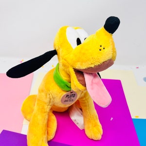 Pu&ograve; includere: Un cane di peluche giallo con orecchie nere e un collare verde. Il cane ha la lingua rosa che sporge e un naso marrone. Il cane &egrave; seduto su una superficie viola e ha un'etichetta che dice "Pluto".