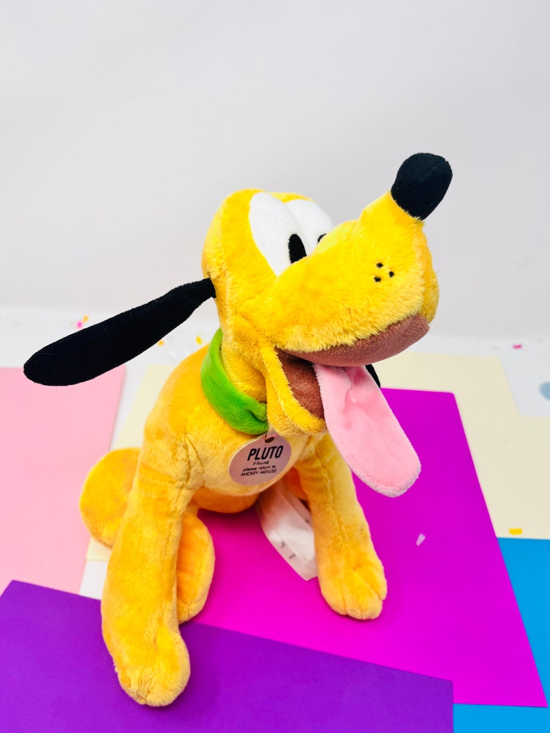 Pu&ograve; includere: Un cane di peluche giallo con orecchie nere e un collare verde. Il cane ha la lingua rosa che sporge e un naso marrone. Il cane &egrave; seduto su una superficie viola e ha un'etichetta che dice "Pluto".