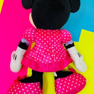 K&ouml;nnte beinhalten: Ein Pl&uuml;schtier von Minnie Mouse, die ein rosa Kleid mit wei&szlig;en Punkten und einer wei&szlig;en Schleife aus Satin tr&auml;gt. Sie hat schwarze Ohren und eine schwarze Nase. Ihre Schuhe sind ebenfalls rosa mit wei&szlig;en Punkten.