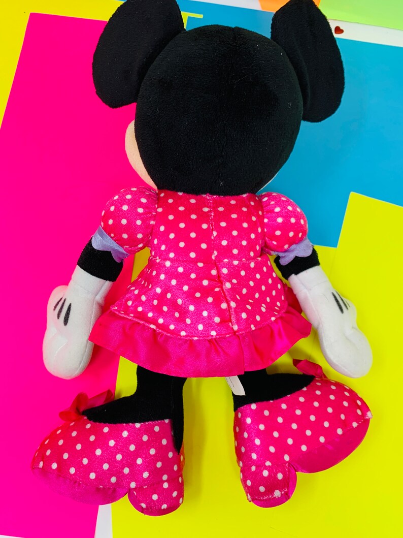K&ouml;nnte beinhalten: Ein Pl&uuml;schtier von Minnie Mouse, die ein rosa Kleid mit wei&szlig;en Punkten und einer wei&szlig;en Schleife aus Satin tr&auml;gt. Sie hat schwarze Ohren und eine schwarze Nase. Ihre Schuhe sind ebenfalls rosa mit wei&szlig;en Punkten.