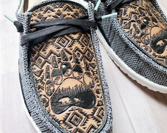 May include: A pair of gray canvas slip-on shoes with a brown leather design featuring a bear paw print and a geometric pattern. The shoes have a white sole and a black lace.