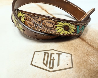 May include: A brown leather belt with a sunflower design and the initials "D&T" embossed on the buckle. The belt is decorated with green and yellow paint.