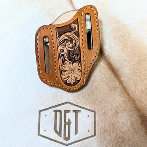 May include: A brown leather knife sheath with intricate floral tooling and the initials "CH" embossed on the front. The sheath is designed to fit a small pocket knife.