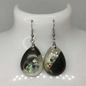 May include: A pair of teardrop-shaped earrings with silver hooks. Each earring features a clear resin section filled with iridescent glitter and a dark, swirled section. The earrings are displayed on a white mannequin head.