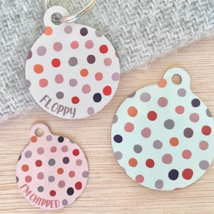 Peut inclure: Trois médailles rondes pour chien avec un motif de pois dans des tons de rose, rouge, gris et orange. Les médailles portent les inscriptions "Floppy" et "I'm chipped".