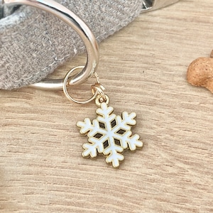 Op de afbeelding: Een gouden sneeuwvlok-bedel met een wit geëmailleerd centrum, bevestigd aan een zilveren ring. De bedel hangt aan een grijs stoffen hondenhalsband.