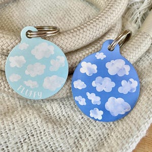 Könnte beinhalten: Zwei runde Haustier-ID-Anhänger mit blauem und hellblauem Hintergrund und weißen Wolkenmotiven. Der Anhänger links trägt die Aufschrift "FLUFFY" in weißer Schrift.