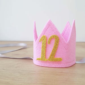May include: Pink felt crown with a gold glitter number 12 on it. The crown has a white satin ribbon attached.