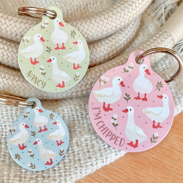 May include: Three round dog tags with white geese and floral designs. The tags are green, pink, and blue. The green tag says "NANCY". The pink tag says "I'M CHIPPED".