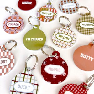 Può includere: Una collezione di medagliette rotonde per animali domestici con anelli metallici. Le medagliette presentano vari motivi, tra cui quadri, tartan e colori a tinta unita. Il testo stampato include nomi come Winnie, Maggie, Cooper, Willow, Teddy, Minnie, Ducky, Dotty e la scritta I'm Chipped.