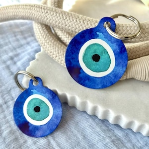 May include: Two round blue keychains with an eye design. The eye has a teal center, a white ring, and a black pupil. The keychains are on a white surface with a cream-colored rope.