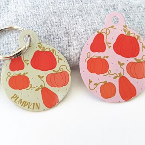 May include: Two round metal pet ID tags with a pumpkin theme. One tag is light green with the word "PUMPKIN" and orange pumpkins. The other tag is light pink with orange pumpkins.