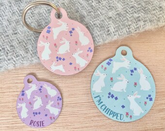 Könnte beinhalten: Drei runde Haustier-ID-Anhänger mit verschiedenen Designs. Der obere Anhänger ist rosa mit weißen Kaninchen und Blumen. Der mittlere Anhänger ist hellblau mit weißen Kaninchen und Blumen. Der untere Anhänger ist lila mit weißen Kaninchen und Blumen. Der untere Anhänger hat das Wort "Rosie" darauf gedruckt. Der mittlere Anhänger hat den Text "I'm chipped." darauf gedruckt.