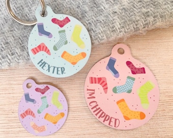 Könnte beinhalten: Drei runde Hundemarken mit bunten Sockenmotiven. Die blaue Marke sagt "Hexter". Die rosa Marke sagt "I'm chipped". Die lila Marke hat eine Vielzahl von bunten Socken.