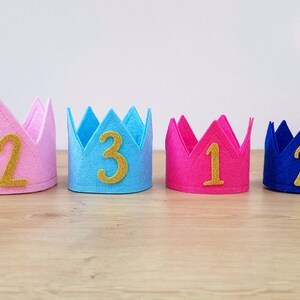 May include: Four felt crowns in different colors, pink, blue, pink, and blue, with gold numbers 12, 3, 1, and 2 on them.