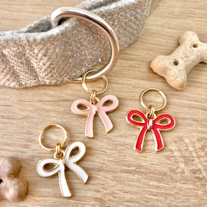 May include: Three gold dog collar charms with different coloured bows. The bows are pink, red and white. The charms are attached to gold rings.