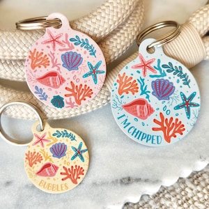 May include: Three round pet ID tags with ocean-themed designs. One tag is pink with a seashell, starfish, and coral design. Another is light blue with the text "I'M CHIPPED". The third is yellow with the text "BUBBLES".