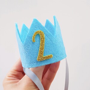 May include: A light blue felt crown with a gold glitter number "2" on the front. The crown has a white ribbon attached to the back.