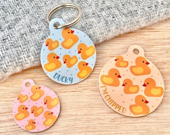 Könnte beinhalten: Drei runde Haustier-ID-Tags mit blauem, rosa und orangefarbenem Hintergrund. Jedes Tag zeigt ein anderes Design mit gelben Gummienenten und dem Text "Ducky" und "I'm chipped".