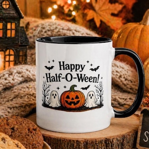 Può includere: Tazza in ceramica bianca con manico e bordo neri, con la scritta "Happy Half-O-Ween!" e una grafica di Halloween con una zucca e dei fantasmi. La tazza è su un sottobicchiere di legno, con biscotti e zucche sullo sfondo.