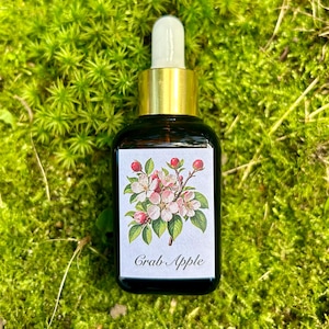 May include: A small glass bottle with a gold cap and a white label. The label has a floral illustration and the text "Crab Apple". The bottle is sitting on a bed of green moss.
