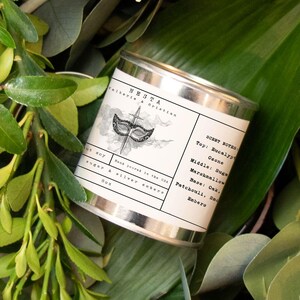 Puede incluir: Una lata de vela plateada con una etiqueta blanca que presenta una ilustraci&oacute;n en blanco y negro de una m&aacute;scara. La etiqueta dice "Home Scent" y enumera las notas de olor:  Salida - Bergamota, Pomelo, Coraz&oacute;n - Jazm&iacute;n, Malvavisco, Fondo - Pachul&iacute;, Cedro, &Aacute;mbar.