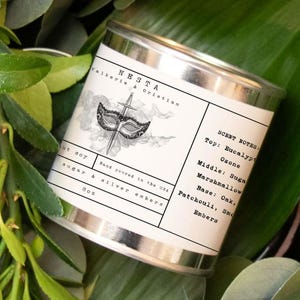 Puede incluir: Una vela en una lata de plata con una etiqueta en blanco y negro que presenta una m&aacute;scara de carnaval y una espada. La etiqueta dice "NESTA Valkerie & Orintin" y enumera las notas de olor: "Top: Eucalipto, Ozono, Middle: Az&uacute;car, Malvavisco, Base: Roble, Pachul&iacute;, Humo, Brasas".