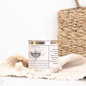 Puede incluir: Una lata de vela plateada con una etiqueta en blanco y negro. La etiqueta presenta una m&aacute;scara de carnaval con una espada y el texto "NESTA A OPIation". Las notas de aroma se enumeran como "Top: Eucal Ozone", "Middle: St Marshmal", "Base: Or Patchouli, Enbera".