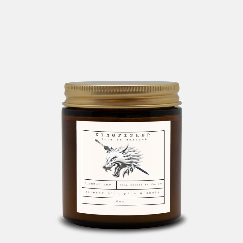 May include: A brown glass jar candle with a gold lid. The label features a wolf illustration with text that reads "KINGFISHER lord of eshitsh". The candle is labeled "coconut soy" and "morning air, pine & smoke".