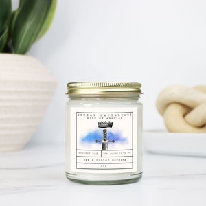 May include: A glass jar candle with a gold lid. The label features text and an illustration of a sword with an eye, a crown, and the words "Dorian Havilliard King of Adarlan". The candle is labeled "sea & winter morning" and is 9 oz.