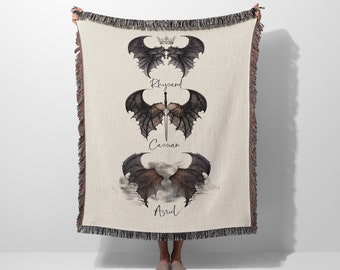 May include: A woven throw blanket with a cream background and three black and brown dragon wings with names "Rhysand", "Cassian", and "Azriel" printed below each set of wings.