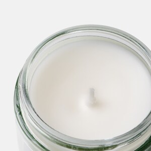 May include: Close-up of a lit candle in a clear glass jar. The candle has a white wax and a single wick. The jar is partially visible, with a clear glass body and a silver rim. The background is a solid white.