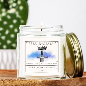 May include: A lit, clear glass jar candle with a gold lid. The label reads "DORIAN HAVILLIARD KING OF ADARLAN" with a crown and sword illustration. The candle is labeled "coconut soy" and "sea & winter morning" and is 4 oz.