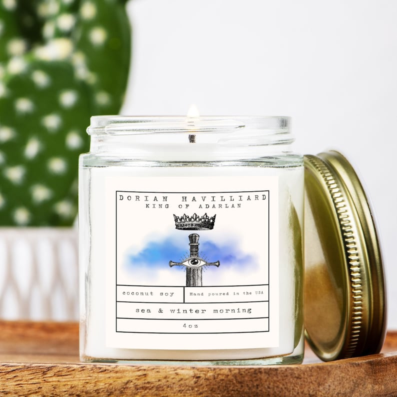 May include: A lit, clear glass jar candle with a gold lid. The label reads "DORIAN HAVILLIARD KING OF ADARLAN" with a crown and sword illustration. The candle is labeled "coconut soy" and "sea & winter morning" and is 4 oz.